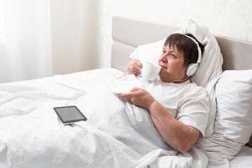 Fototapeta premium Morning coffee for grandma. An elderly woman with headphones listens to music and drinks coffee.