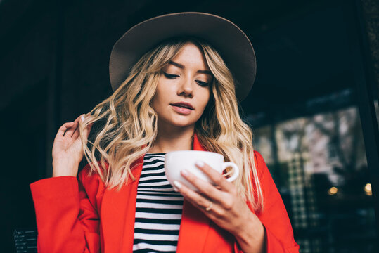 Beautiful hipster girl in stylish hat spending leisure pastime in street cafe holding white cup with hot caffeine beverage and thinking about divination on coffee grounds, Caucasian female customer