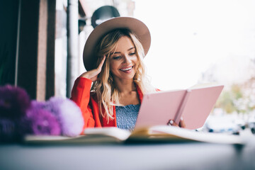 Cheerful hipster girl in stylish hat reading literature book during free time in street cafe, happy Caucaisn student checking education information in textbook while learning and smiling outdoors