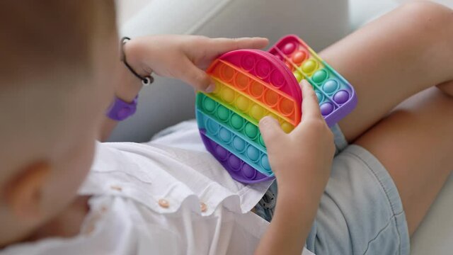 Portrait blonde boy with pop it sensory toy different forms. Boy presses colorful rainbow squishy soft silicone bubbles in children's playroom. Stress and anxiety relief. Trendy fidgeting game.