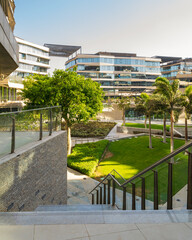 Marble staircase and glass handrail leading to street in contemporary urban city district with well run glass buildings, verdant lawn, and palm trees on sunny day