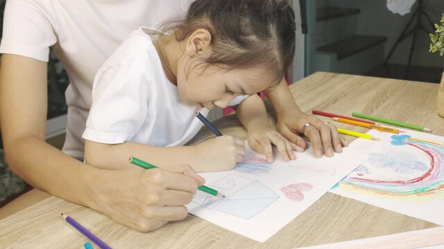 Adorable asian kid girl is drawing and coloring beautiful and colorful pictures (rainbow, house) with her mother together shows concept of happy parental leisure time activity with family.