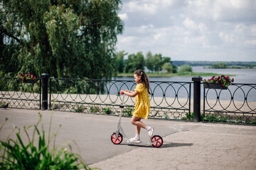 lifestyle portrait a six-year-old girl in a yellow dress rides a scooter along the river on a sunny day with green trees, grass on the background. 