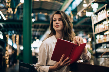 Contemplative hipster girl in casual clothes holding literature best seller in hands and thinking...