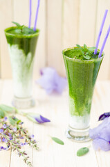 Two glasses of Healthy green smoothie with spinach, kiwi and oranges on light wooden table. Flowers in the background.