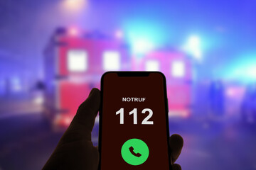 Person hält Smartphone in der Hand und ruft den Notruf 112, im Hintergrund Rettungswagen und...