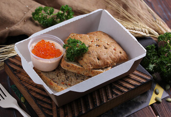 Healthy food for delivery. Breakfast. Boiled eggs with red caviar, crackers and yellow sauce and herbs in a white lunch box on a wooden board on a dark gray background. Rustic. Background image