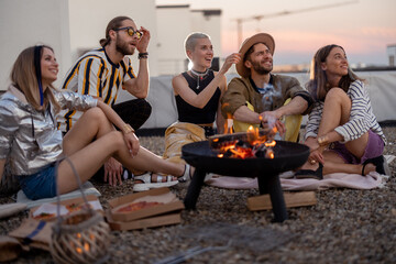 Group of young stylish friends sitting together by the fireplace, having a picnic on the rooftop terrace at dusk. Enjoy summertime and beautiful sunset view