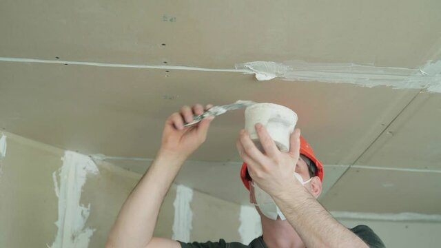 young man in a builder's uniform puts putty on the plasterboard ceiling. finishing the joints of drywall sheets with putty. 4k video resolution
