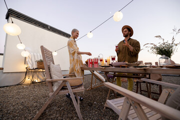 Young stylish friends hang out together talk and having fun on a the beautifully decorated rooftop terrace at dusk. Couple at open air party, wide view