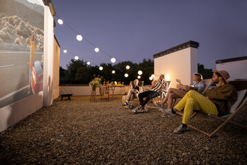 Small group of people watching movie on the rooftop terrace at sunset. Open air cinema concept. Romantic leisure and entertainment on the roof of a country house