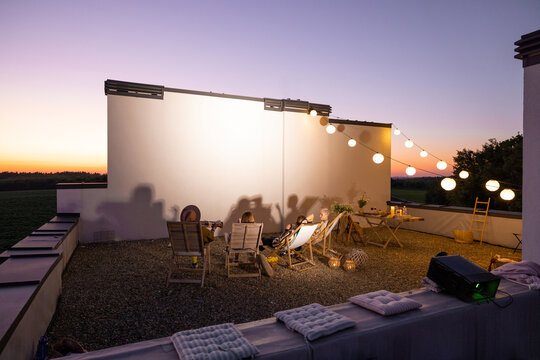 Small Group Of People Watching Movie On The Rooftop Terrace At Sunset. Open Air Cinema Concept. White Projection On The Wall With Copy Space