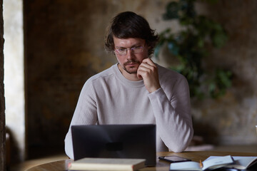 Concentrated serious young man working from home and enjoying distant job. male student using laptop for internet learning and creative writing and dreaming.