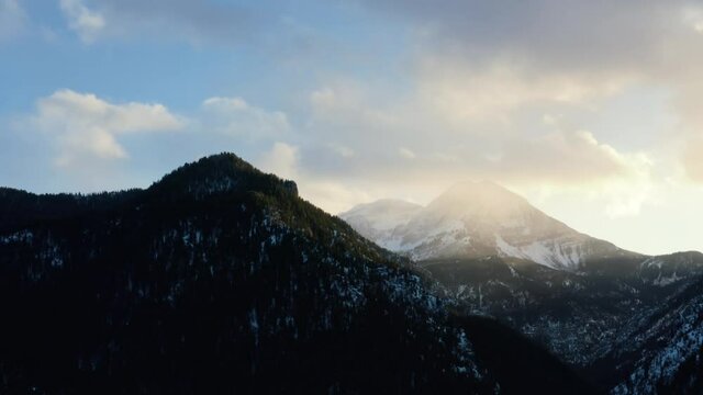 Aerial drone shot of a winter landscape of Mount Timpanogos in the background surrounded by a pine tree forest during sunset from the frozen Tibble Fork Reservoir in American Fork Canyon, Utah
