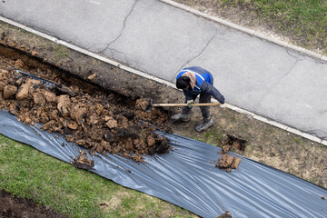 A worker digging a ditch along a pavement in summer.