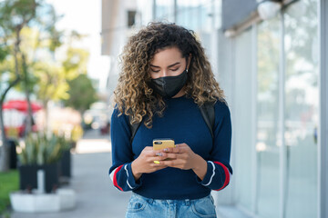 Latin woman wearing a face mask while using her mobile phone outdoors.