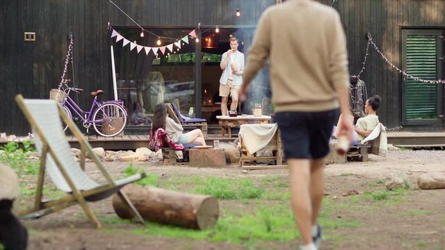 Follow shot of young man with beer bottle joining diverse group of friends talking and having fun at outdoor summer barbecue party in backyard. Young people gathering together again after restrictions