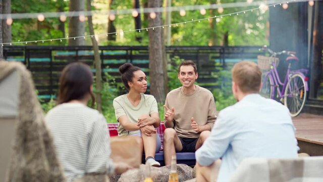 Tilt down of group of four diverse young friends, two couples, sitting under lights around fire, talking joyfully and laughing at double date. Man telling funny story to friends