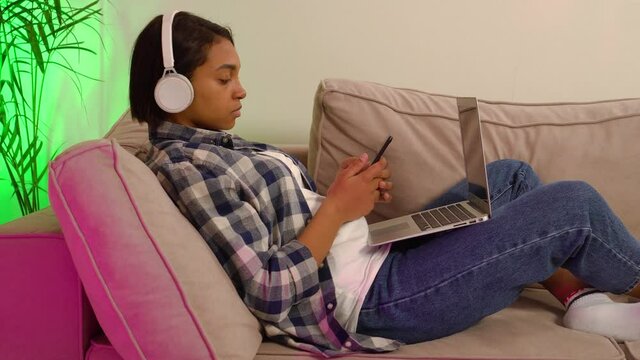 An African-American woman lying at home on the couch works online using a laptop and smartphone. He listens to music with big white headphones. The concept of working online and freelancing.