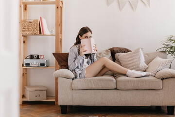 Cute woman in pajamas and socks is resting on sofa and covers her face with book. Pretty young lady in home outfit holds pink notebook and sits on beige couch