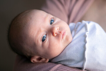 Close-up of a newborn baby with no hair and blue eyes, lying on the mother's arm and looking with his head turned. The child is wrapped in white clothes. Head turn.