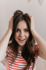 Girl with green eyes looks into camera and ruffles her wavy hair. Brunette lady in striped shirt happily posing on white background