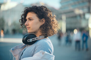 A young woman standing on a street with earphones around neck and looking forward..