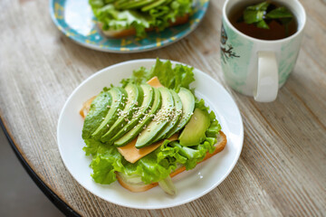 Avocado toast on wheat bread with cheese, salad and sesame seeds on a white plate on a wooden table. Healthy breakfast.