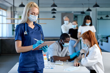 Female doctor at work, beginning of worldwide mass vaccination for coronavirus COVID-19, influenza or flu, world immunization concept. Portrait of blonde nurse in blue uniform holding tablet, writing