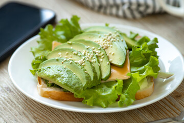Avocado toast on wheat bread with cheese, salad and sesame seeds on a white plate on a wooden table. Healthy breakfast.