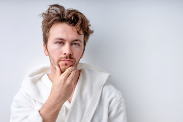 Calm young man admiring touching face, looking at camera confidently, wearing bathrobe, Caucasian guy in the morning. Poeple lifestyle, morning routine, leisure concept. Copy space. Beauty concept
