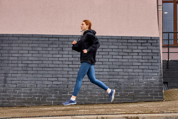 Side view on caucasian sportswoman jogging running in the morning, in urban area, wearing black hoodie and blue leggins, concentrated on sport training, Healthy lifestyle, sport, fitness concept
