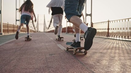 Close-up of the legs of unrecognizable skateboarders riding across the bridge at dawn. Group of skaters, boys and girls, ride their skateboards - Powered by Adobe