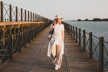 A girl in light clothes and a hat is walking along the pier and looking at the sunset against the backdrop of the seascape. Huelva, Spain.