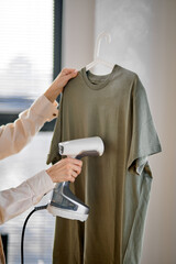 Cropped woman using steaming iron to ironing fashion shirt at home in bright cozy room. Female holding electric steamer, using modern technologies. Copy space. Side view © Roman