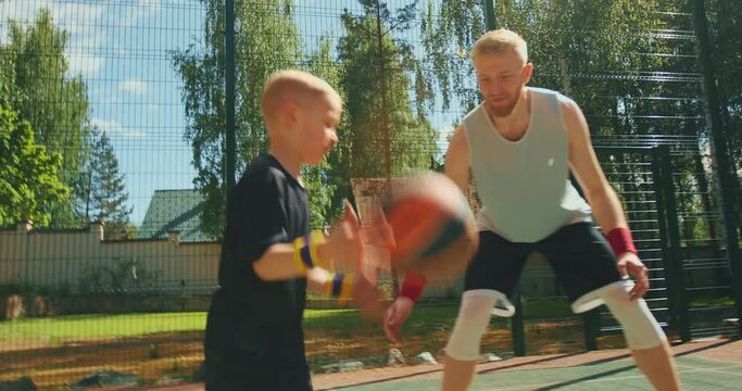 Basketball player sportive guy training child boy to dribble on basketball court outdoors in summertime