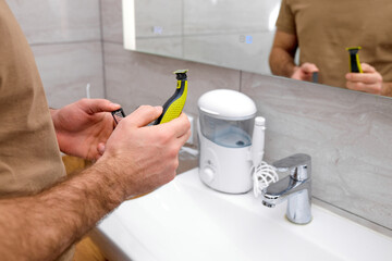 cropped man is going to shave beard with electric shaver, close-up photo of hands, at home bathroom. side view on caucasian guy taking care of face skin and hair. copy space. smart devices