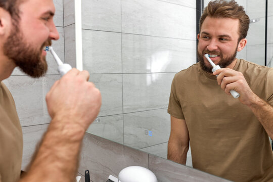 Young Caucasian Man Brushing Teeth With Electric Toothbrush During Morning Hygiene Procedures In Bathroom, At Home. Handsome Bearded Guy In Casual T-shirt Look At Mirror Reflection, Morning Routine