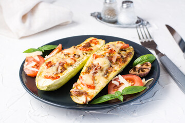 Ready-to-eat baked zucchini halves filled with cheese and tomato and basil leaves on a black plate on a white table. Vegetable menu, healthy food