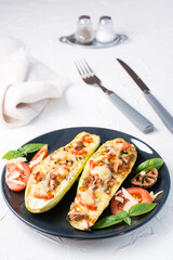 Ready-to-eat baked zucchini halves filled with cheese and tomato and basil leaves and cutlery on a black plate on a white table. Vegetable menu, healthy food. Vertical view