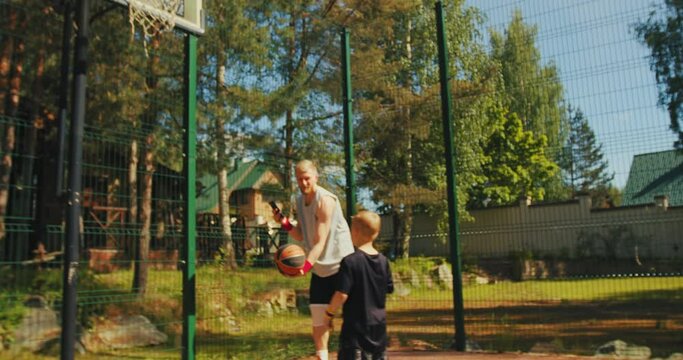 Little sportive kid boy throwing ball in basketball hoop training outdoors, father take video on phone camera