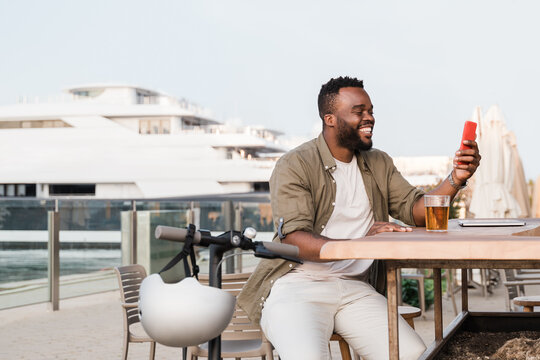 Young african american man doing video call on smartphone outdoor at brewery bar restaurant while drinking beer - Focus on face