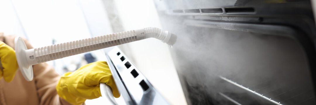 Woman in protective gloves washing oven with steam brush closeup