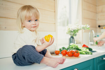 Baby cute blonde boy playing with vegetables in the kitchen. Waiting for your lunch of seasonal summer products. Comfortable children's home clothes.
