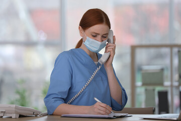 Receptionist with protective mask talking on phone at countertop in hospital