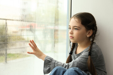 Sad little girl near window indoors on rainy day