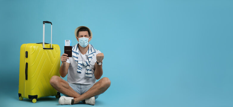 Male tourist in protective mask holding passport with ticket near suitcase on turquoise background