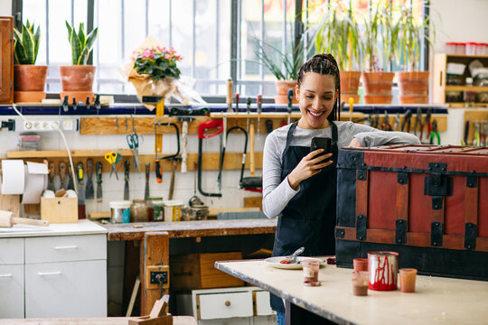 Cheerful craftswoman with smartphone in woodwork workshop 