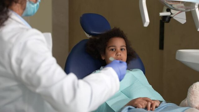 Scared curly child sitting on stomatological chair while female dentist examining her teeth before treatment. Little preadolescent girl opening mouth for stomatologist in children's dental clinic