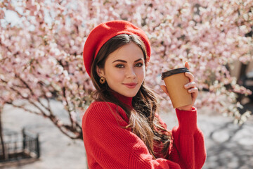 Beautiful girl in beret and stylish earrings holding glass of coffee on background of sakura....